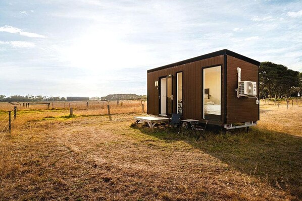Uma cabana pequena, com um único quarto, porta que dá para uma área de dormir, uma mesinha e uma cadeira lá fora.