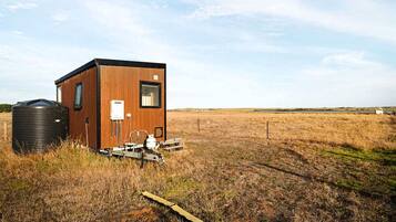 Une petite cabane en bois, équipée d’un réservoir d’eau et d’un système électrique, se trouve dans un champ herbeux.