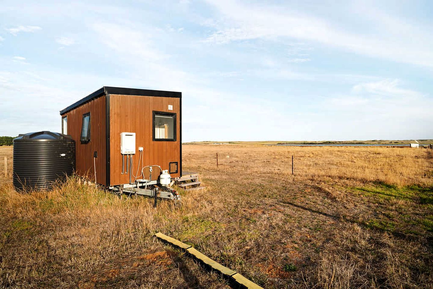 Une petite cabane en bois, équipée d’un réservoir d’eau et d’un système électrique, se trouve dans un champ herbeux.