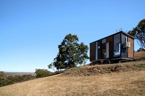 Una cabana moderna d'un pis, amb una finestra gran, situada en un turó verd amb cel blau.
