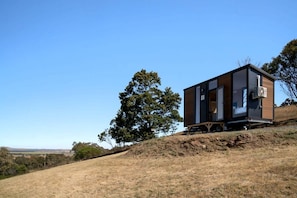 A modern, single-story cabin with a large window, situated on a grassy hill with a clear sky.