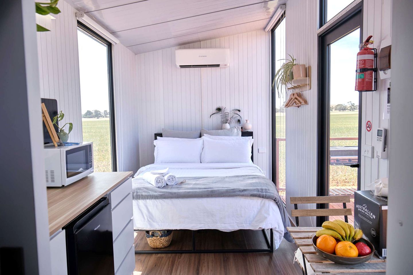 Ein kleines Zimmer mit Bett, Mikrowelle, Tisch, Feuerlöscher und Fenster mit Blick ins Freie.