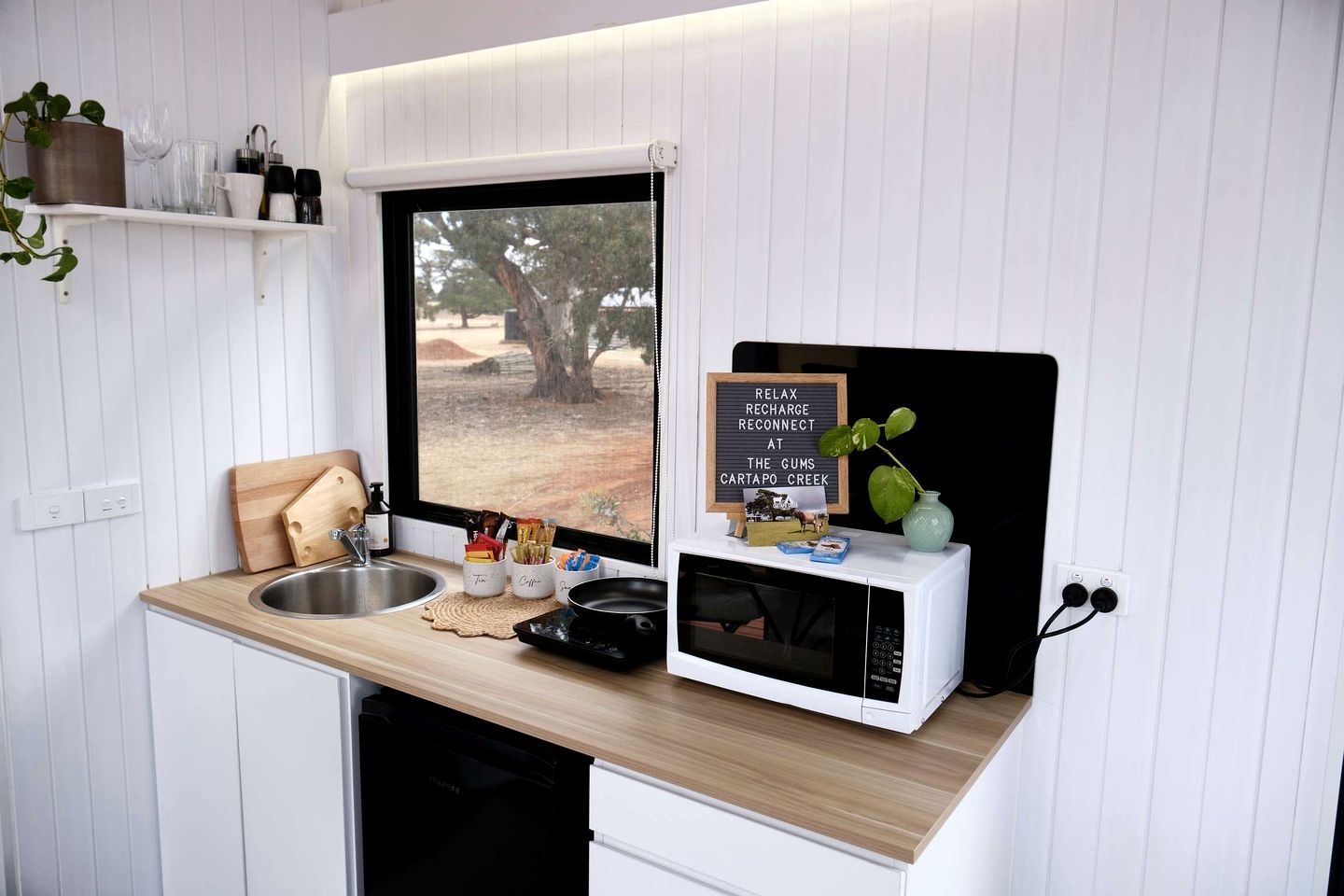 Uma cozinha compacta com janela, pia, micro-ondas e um pequeno balcão com diversos objetos.