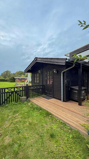 Een houten vakantiehuis met een veranda, een kleine tuin en een rood gebouw op de achtergrond.