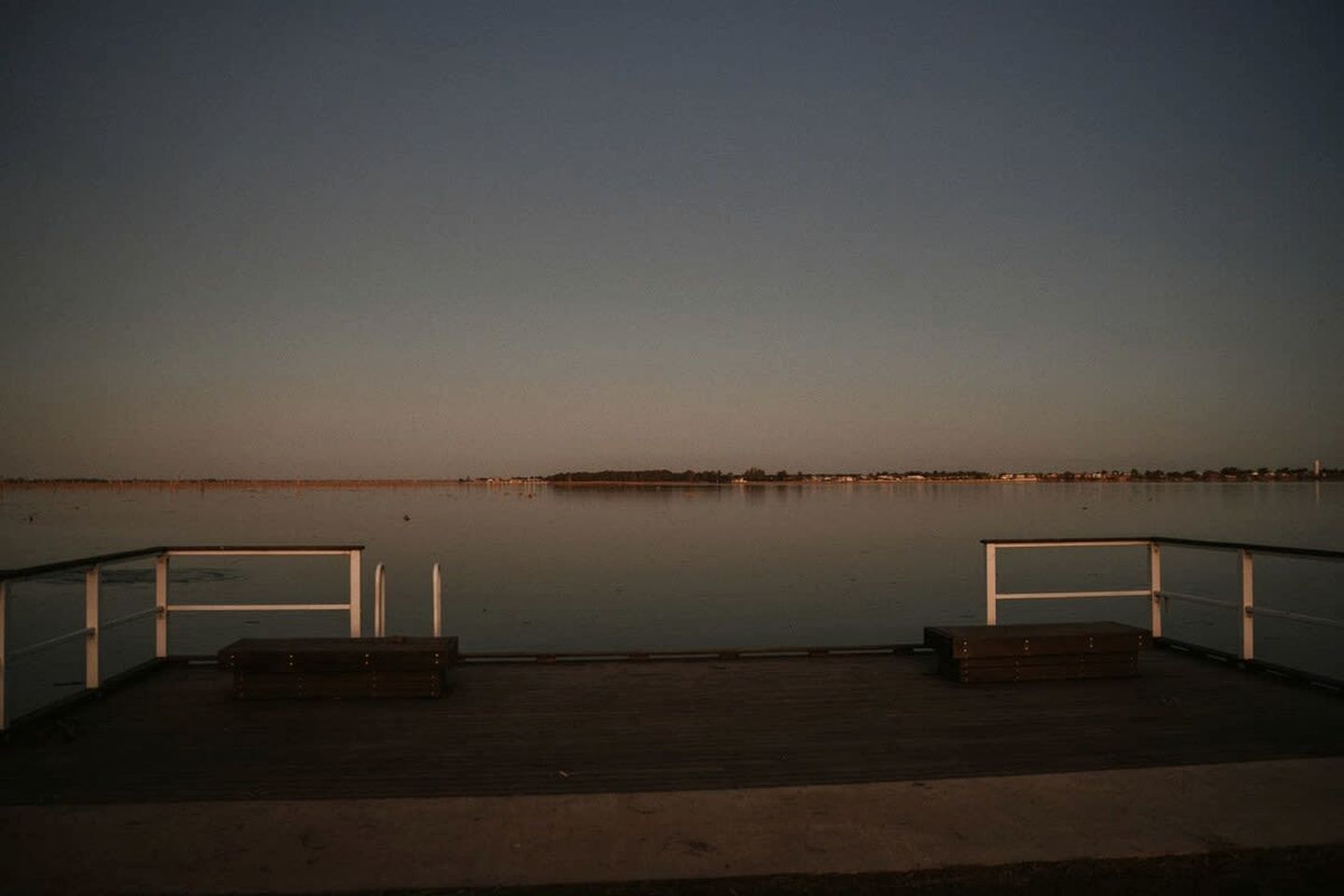 A wooden pier with railings extending over calm water, leading to a distant shoreline.
