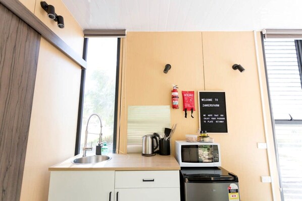 A compact kitchenette with a sink, microwave, and a small stove. There is a fire extinguisher and a welcome sign on the wall.