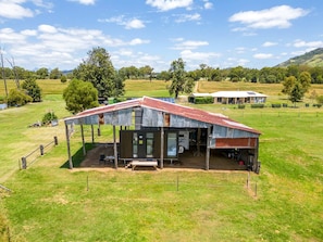 A rustic metal-roofed building with a covered outdoor seating area, surrounded by a grassy field and trees.