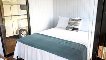 Un lit avec une literie blanche et un plaid turquoise, un plancher en bois et une fenêtre donnant sur une caravane.