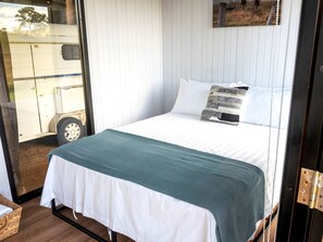 A bed with white bedding and a teal throw, a wooden floor, and a window with a view of a trailer.