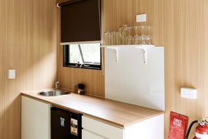 Een compact keukenhoekje met een spoelbak, een raam met zonwering, een koelkast en een brandblusser.