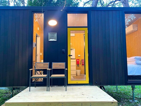 Eine kleine Holzhütte mit einer Holzterrasse, zwei Stühlen, einer Tür mit gelbem Rahmen und einem sichtbaren Innenraum mit einem Bett und einem roten Feuerlöscher.