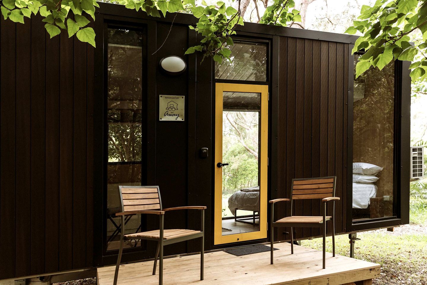 Uma pequena cabana de madeira com porta, duas cadeiras de madeira e um banco lá fora.