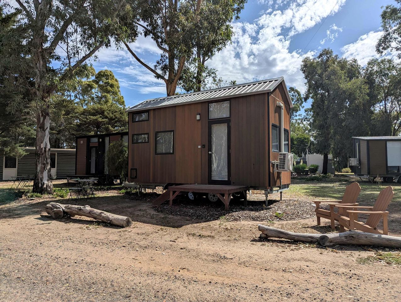 A small wooden cabin with a door and windows, situated on a gravel ground with trees in the background.