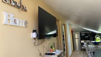 Ein modernes Wohnzimmer mit einem wandmontierten Flachbildfernseher, einem Essbereich mit gestreiftem Tischläufer und einer Küchenzeile mit Edelstahl-Kühlschrank.