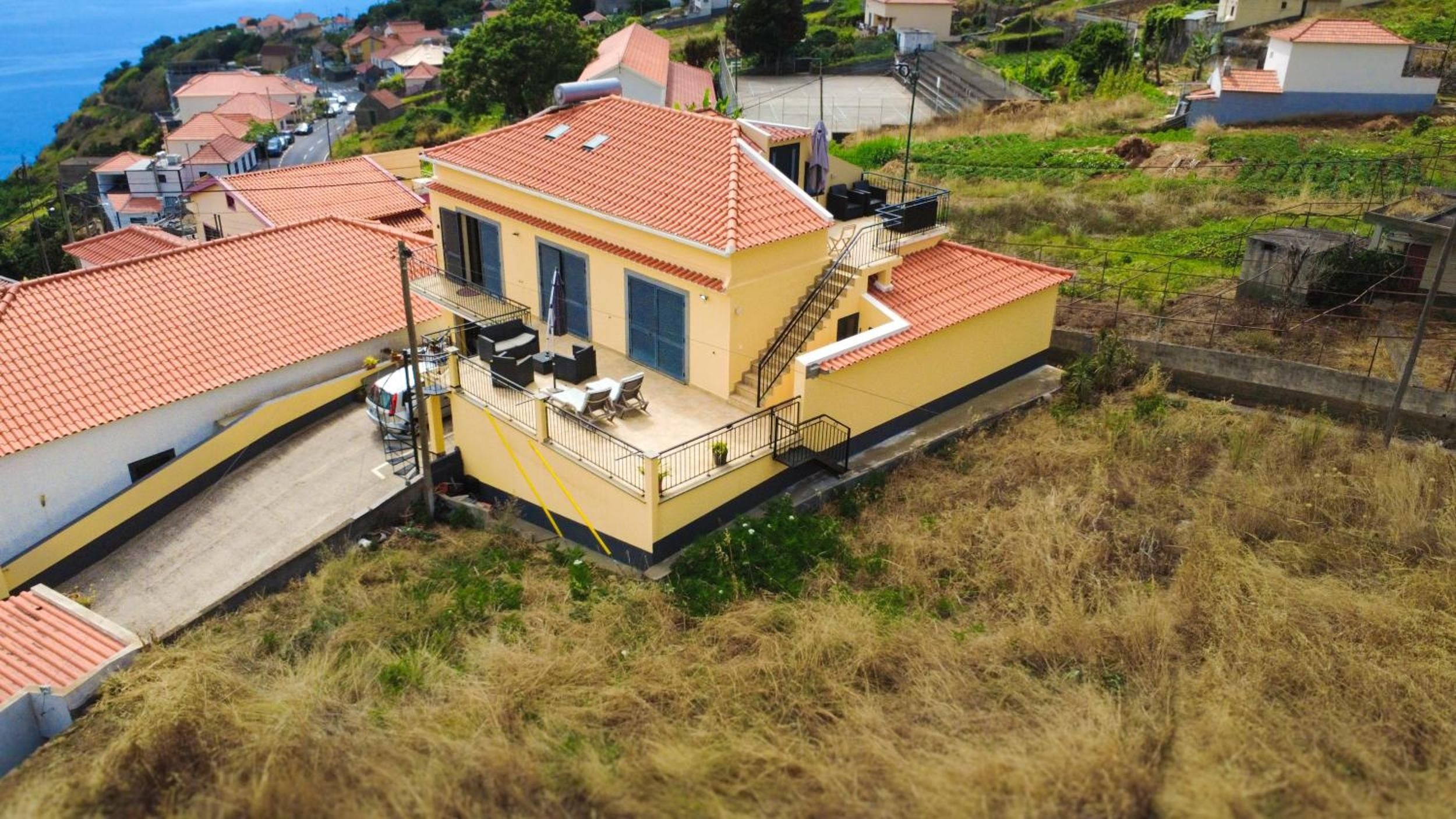 Um edifício de dois andares com telhado de telhas vermelhas, uma varanda com móveis para área externa e vista para o mar.