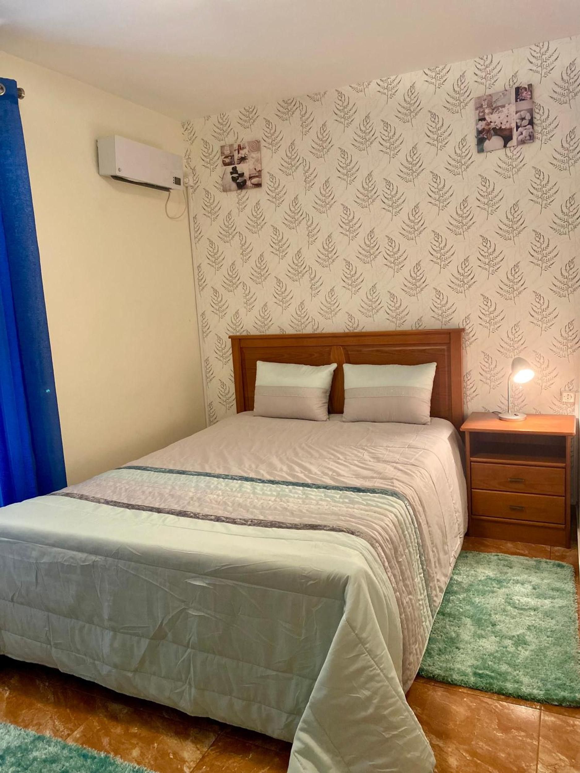 A bedroom with a bed, a nightstand, a lamp, a wall air conditioner, and a patterned wallpaper.
