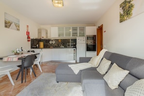 A modern living room with a grey sofa, a dining area with a table and chairs, and a kitchen with white cabinets and appliances.
