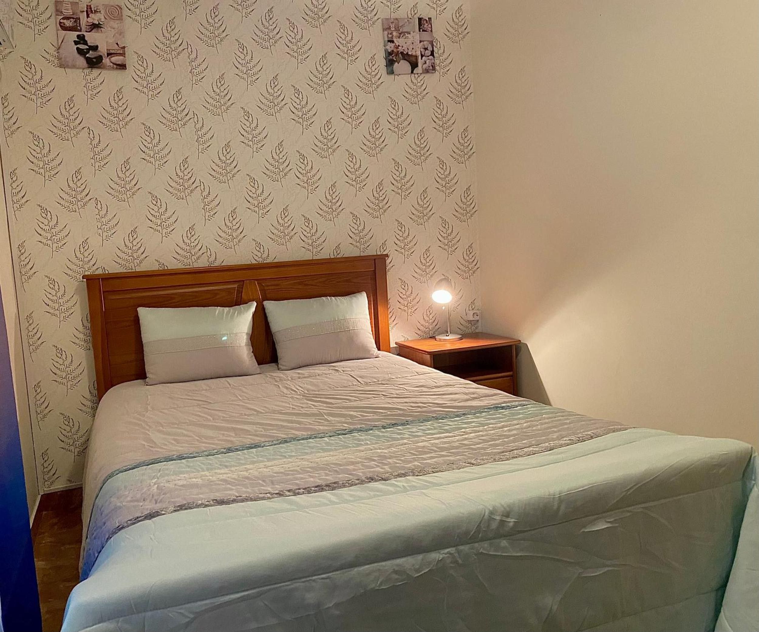 Uma cama de solteiro com cabeceira de madeira, duas almofadas, mesa de cabeceira com abajur e parede com papel de parede com tema da natureza.