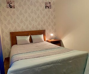 Una cama individual con cabecera de madera, dos almohadas, una mesita de noche con lámpara y un papel tapiz con motivos naturales.