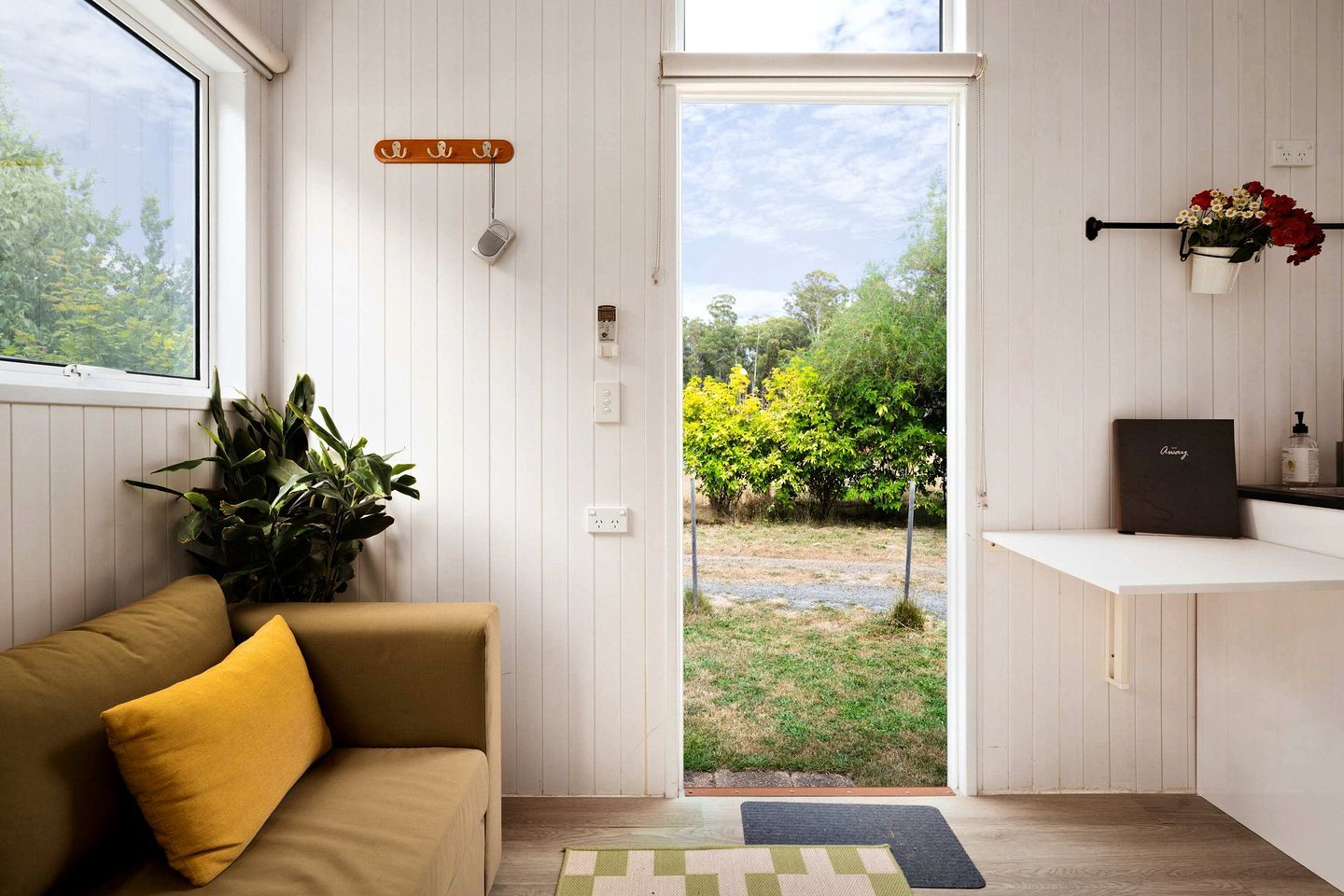 A room with a sofa, a potted plant, a door leading outside, and a small shelf with a book and a bottle.