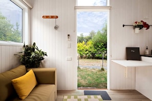 A room with a sofa, a potted plant, a door leading outside, and a small shelf with a book and a bottle.