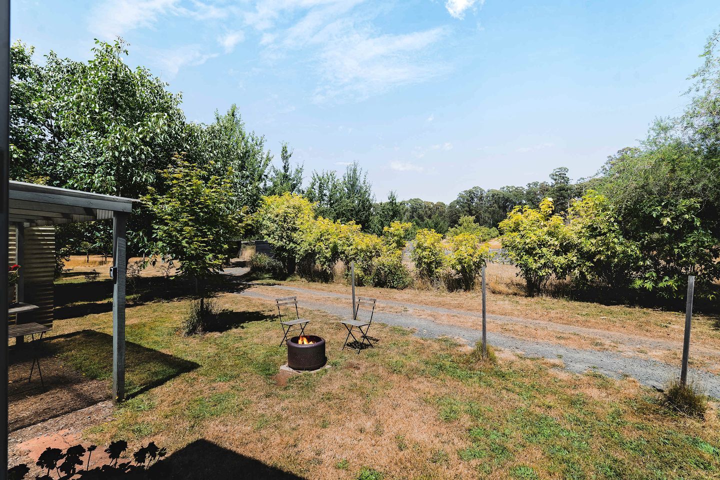 A covered outdoor seating area with a fire pit and chairs, surrounded by trees and a gravel path.