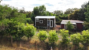 A small, single-story wooden house with a metal roof, surrounded by dense greenery and a fenced yard.