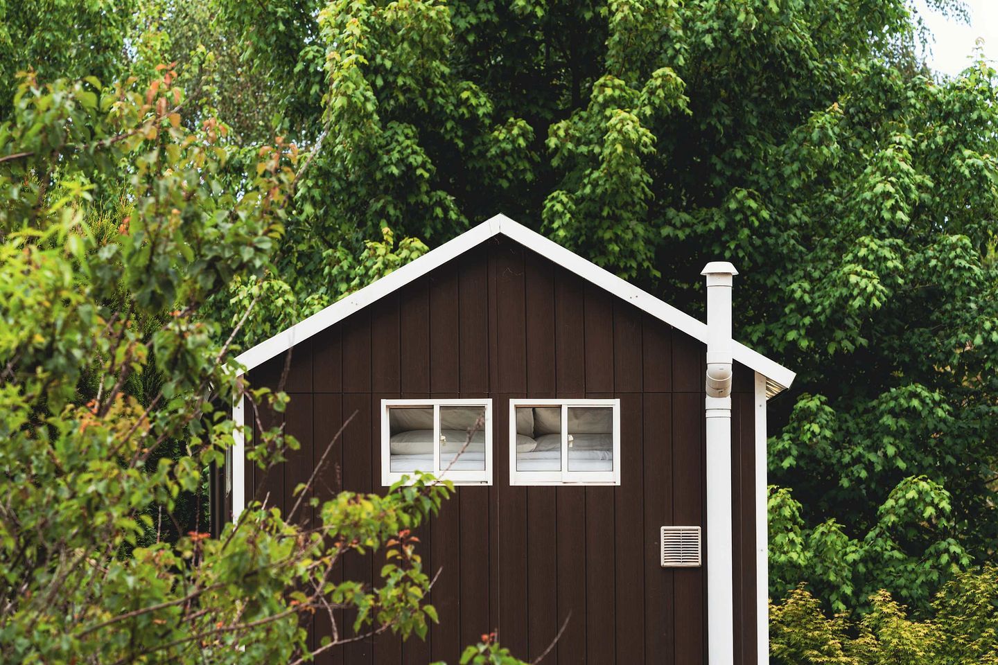A small, dark wooden cabin with two windows and a chimney, surrounded by dense foliage.