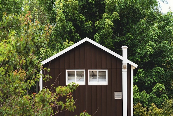 A small, dark wooden cabin with two windows and a chimney, surrounded by dense foliage.