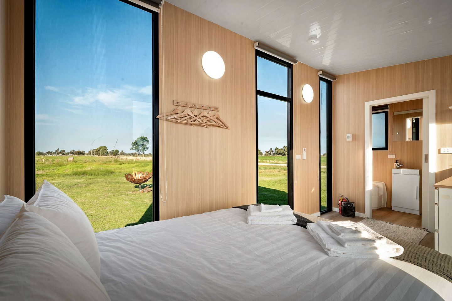 A modern bedroom with a large bed, a view of a grassy field, and a small kitchenette.