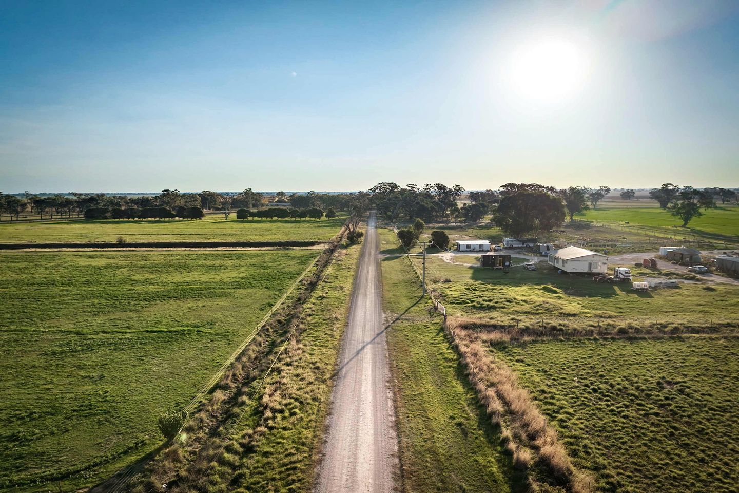 Et landligt landskab med en lige vej, der fører til en gruppe bygninger, omgivet af åbne marker og træer.