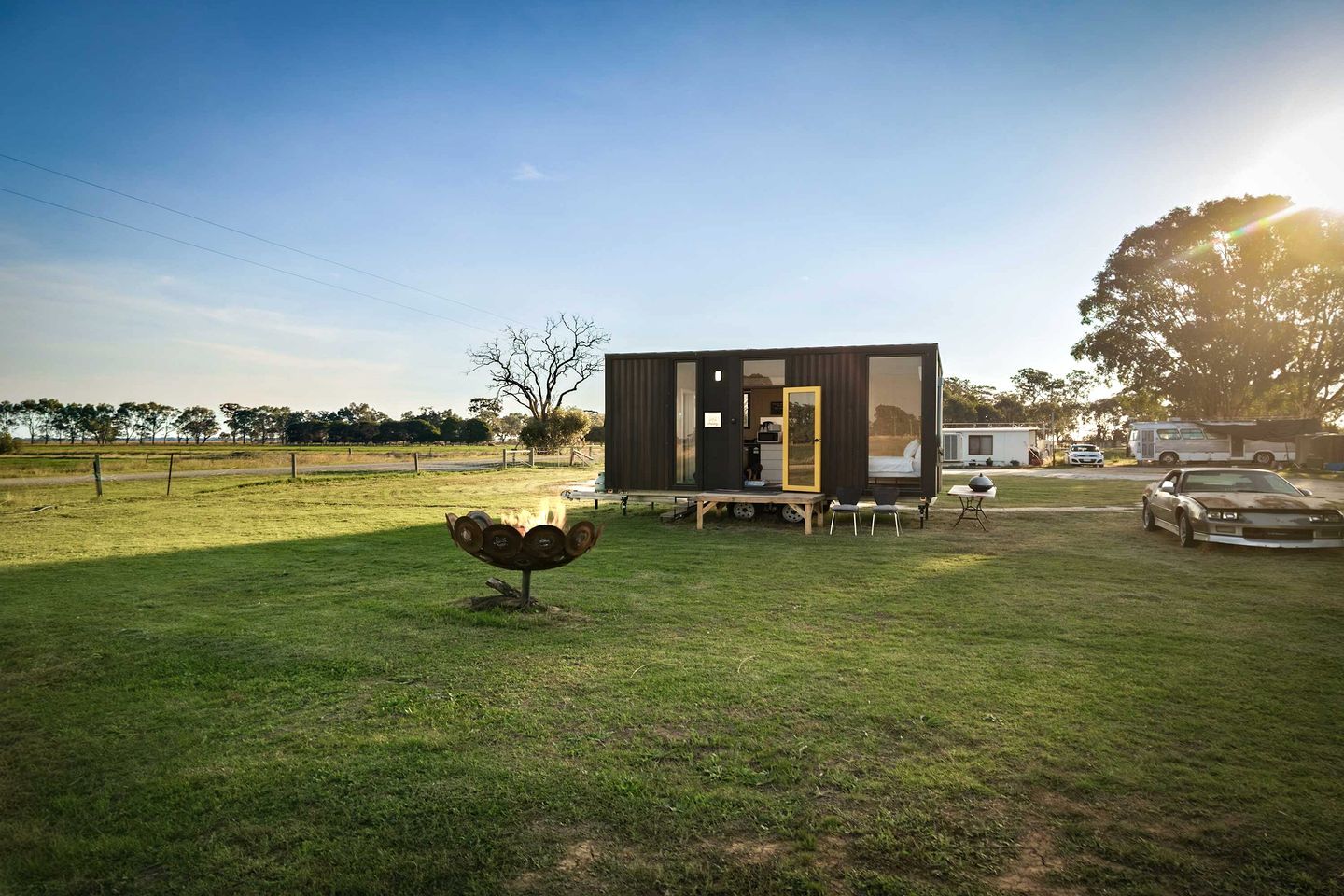 A modern, compact mobile home with a large sliding door, a small deck, and a fire pit in the yard.