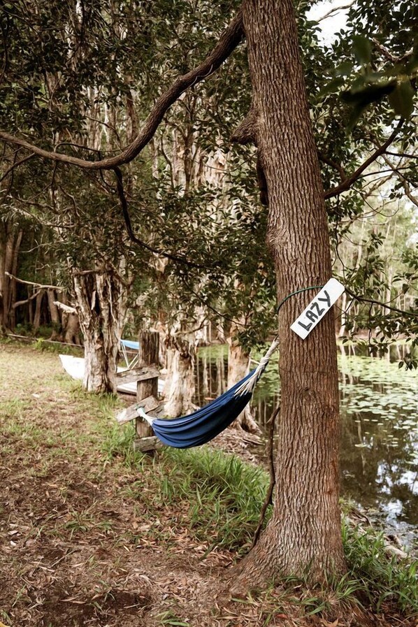 Un hamac est attaché à un arbre, et une pancarte indique le mot « PARESSE » dans une zone boisée, près d’une étendue d’eau.