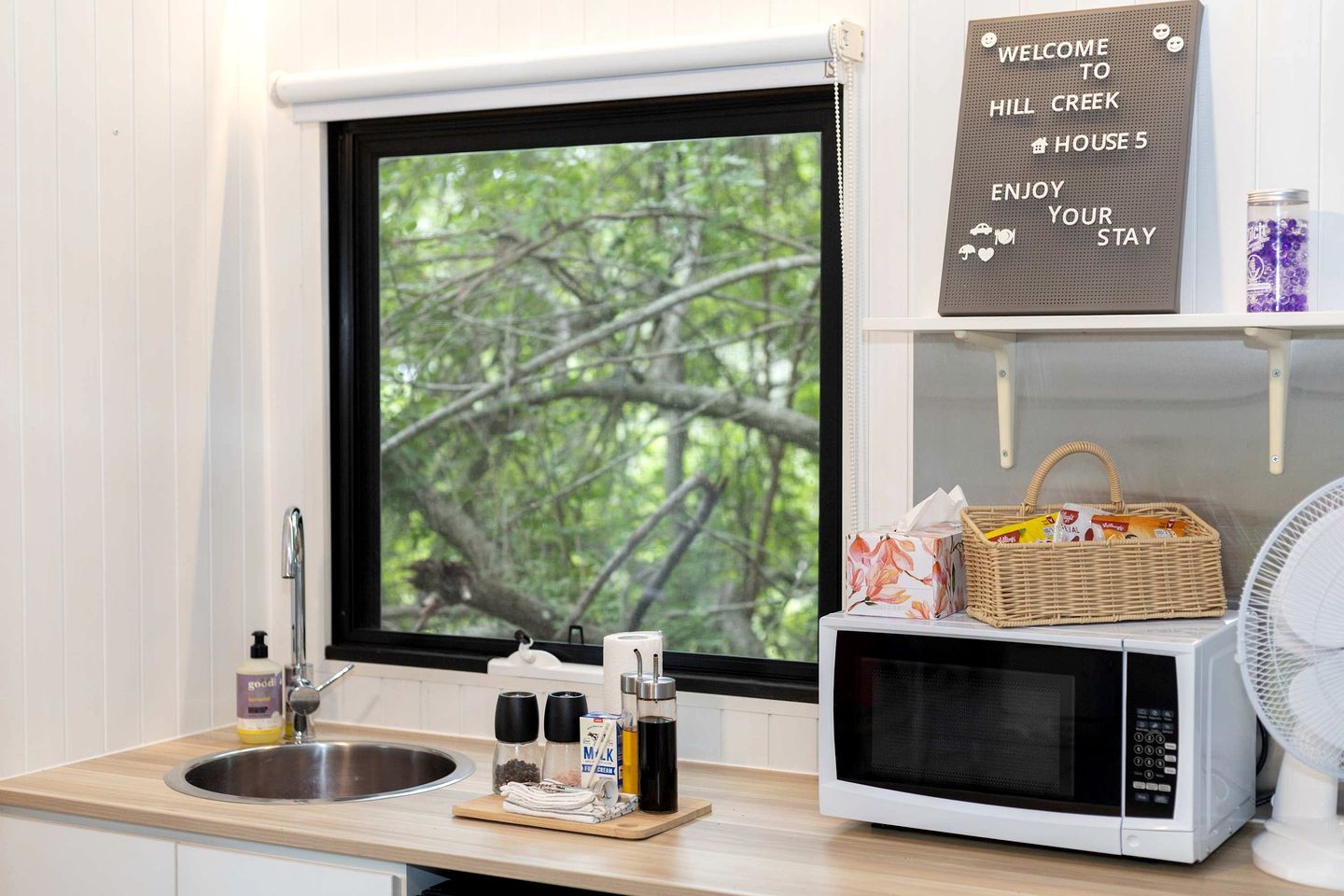 A kitchen with a sink, a microwave, and a window with a view of trees. There's a welcome sign above the window and a basket with snacks on the counter.