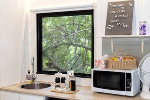 A kitchen with a sink, a microwave, and a window with a view of trees. There's a welcome sign above the window and a basket with snacks on the counter.