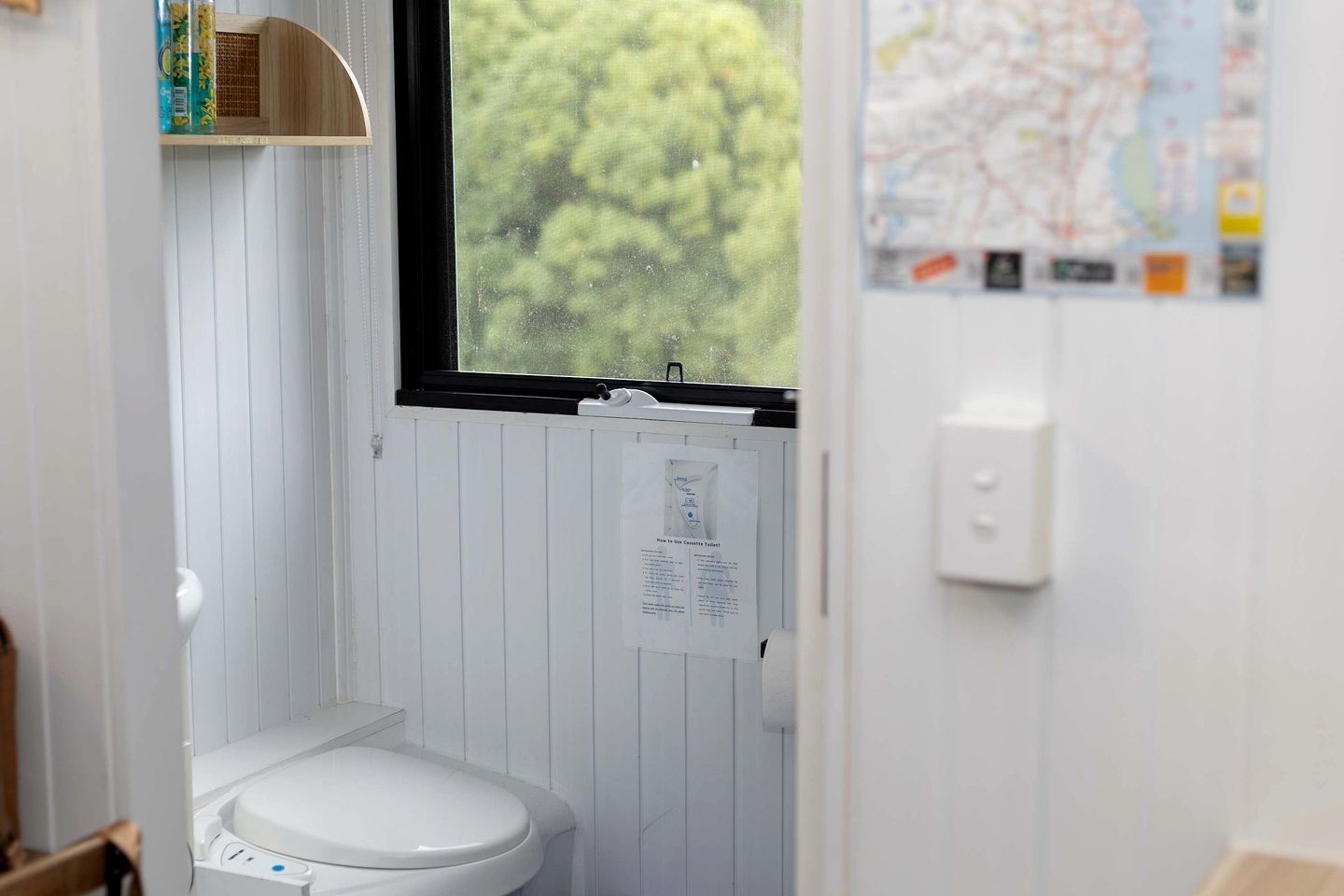 Une petite salle de bain blanche, avec des toilettes, une fenêtre et une carte accrochée au mur.