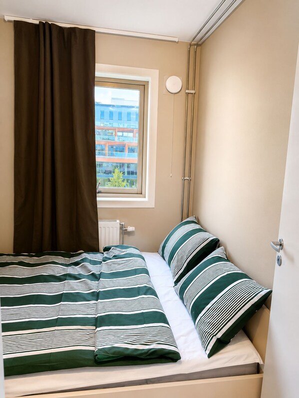Ein Einzelbett mit gestreifter Bettwäsche, ein Fenster mit Blick auf Gebäude, eine Heizung und eine Tür mit Griff.