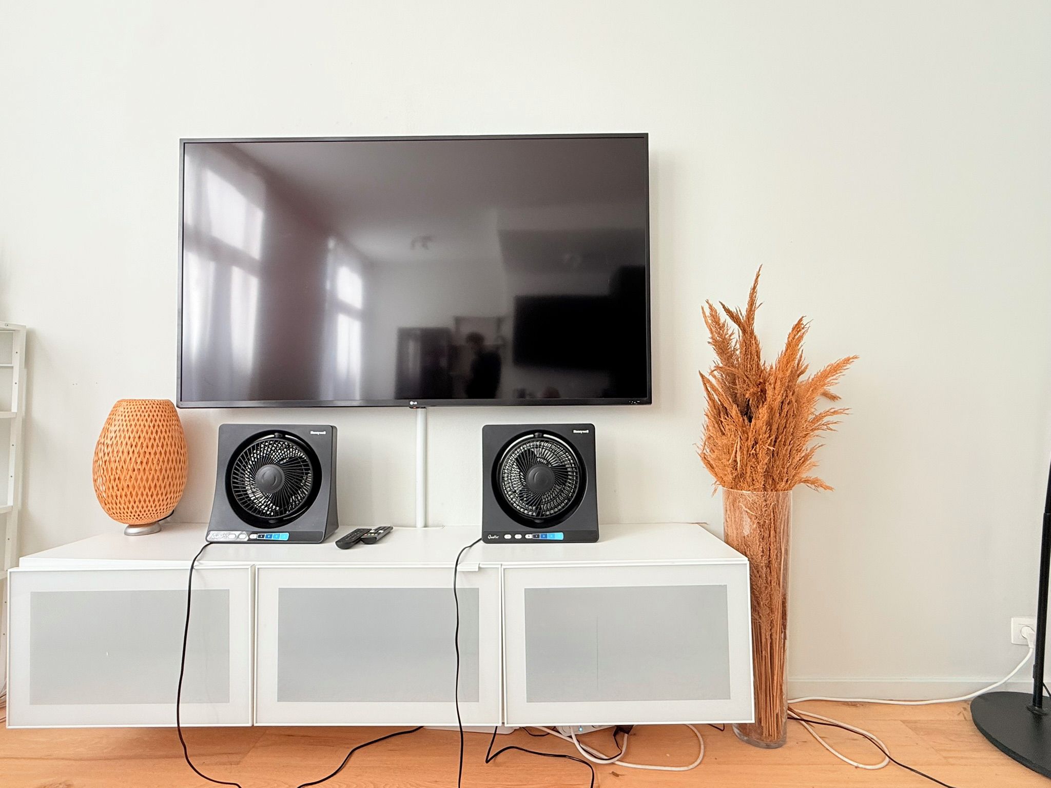 Televisão de tela plana presa na parede acima de um móvel branco com dois alto-falantes pretos e um vaso decorativo.
