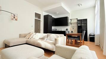 A modern living room with a sectional sofa, a dining area with a wooden table and chairs, and a kitchen with black cabinets and a microwave.