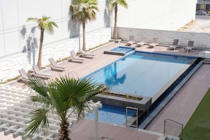 Ein rechteckiger Swimmingpool mit einer gefliesten Kante, umgeben von Liegestühlen und Palmen.