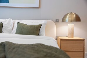 Ein ordentlich bezogenes Bett mit weißen und grünen Kissen, einer Nachttischlampe und einem Holfnachtkästchen.