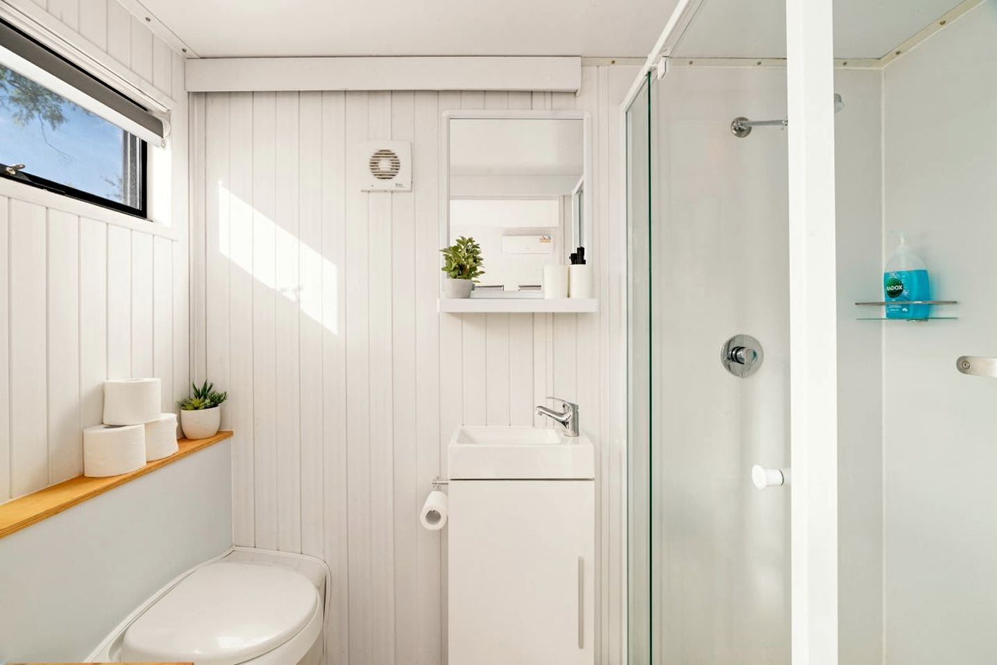 A bathroom with a toilet, sink, and shower. There is a shelf with toiletries and a potted plant.