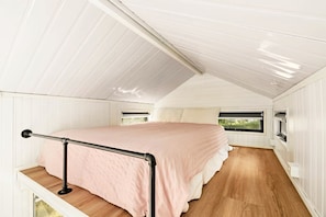 A bedroom with a bed, wooden flooring, white walls, and a window.