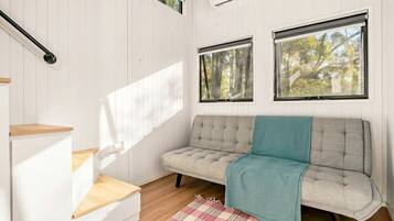 Una habitación con un sofá, una mesa de madera, un tapete escocés y dos ventanas con vistas a árboles.