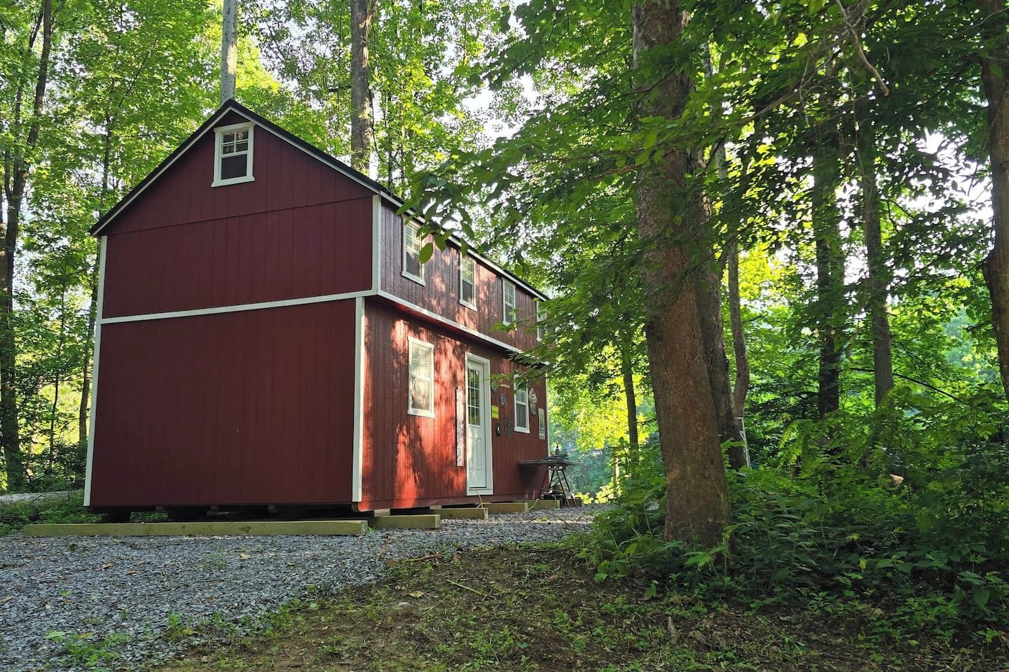 A small, single-story red building with a white door and windows, surrounded by trees and gravel.
