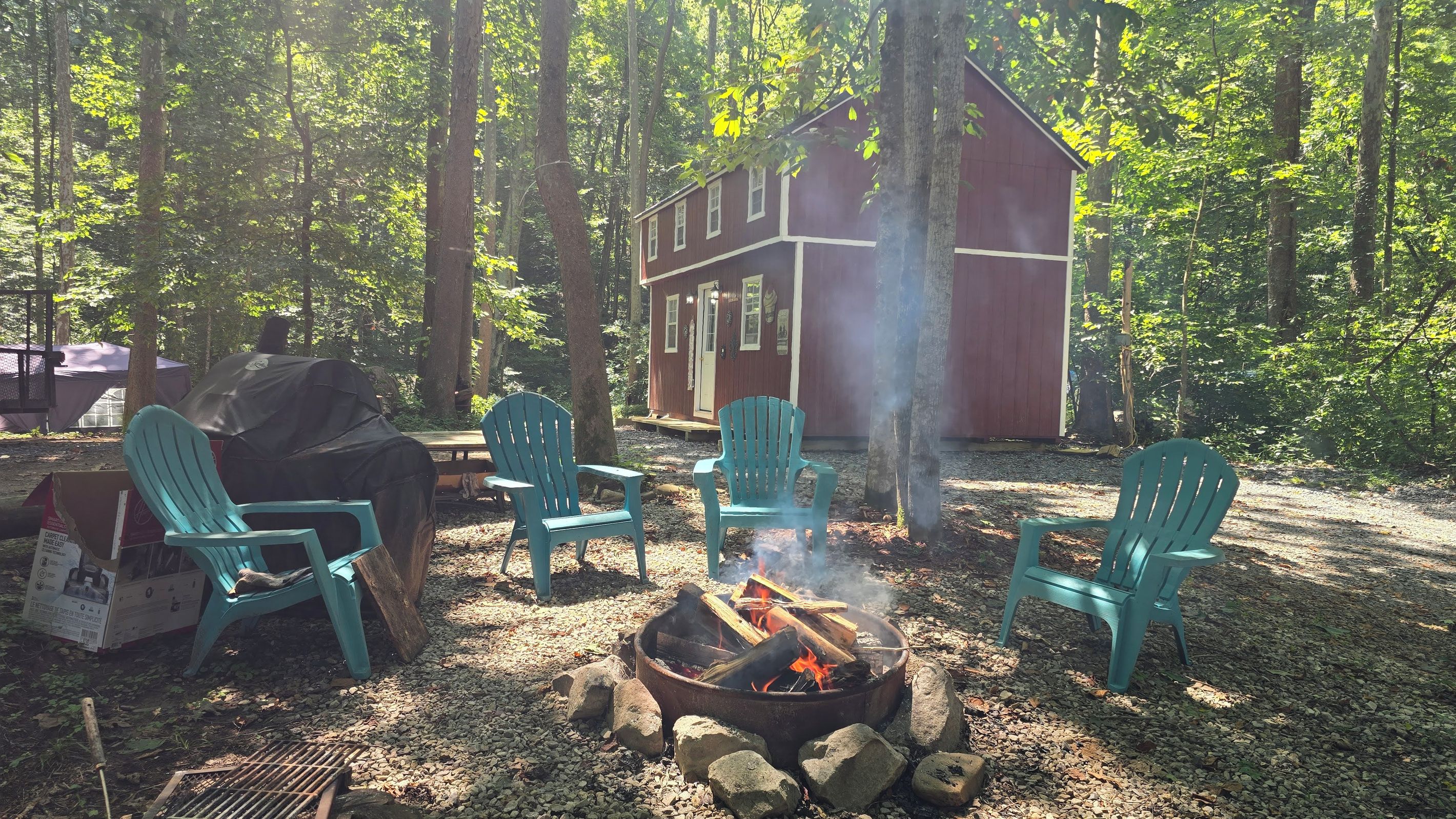 A forested area with a red building, a fire pit with logs, and turquoise chairs.