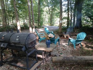 Un espacio al aire libre con barbacoa, una parrilla, sillas y una mesa en zona boscosa.