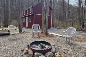 A red building with a fire pit and three white plastic chairs in a forested area.