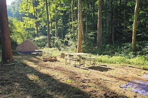 Un camping en el bosque con una tienda de campaña, mesas de picnic y un lomo azul en el suelo.