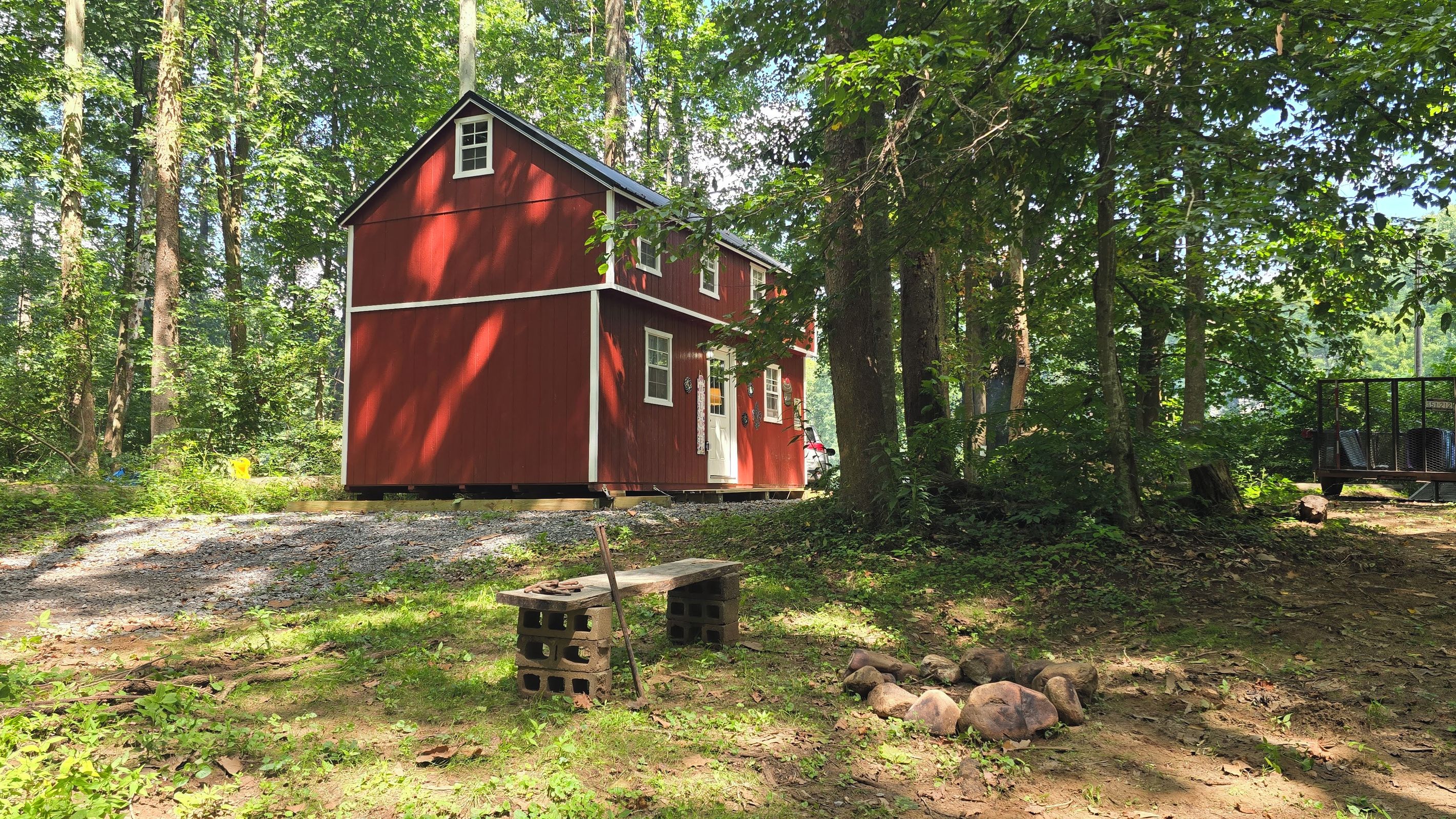 Une maison en bois rouge, dotée d’un petit porche et d’une fenêtre, entourée d’arbres et bordée d’un chemin de gravier.